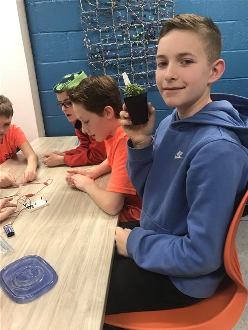Student hold up plant to show the sensor that he built that determines if the plant needs water.  Students in the background. 